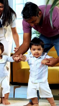Indian Asian young parents helping twin infant babies walk by holding their hands inside modern home living room, guiding first steps together as smiling babies enjoy playful family bonding moment