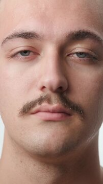 Frontal studio close-up portrait of a sweating young Caucasian man with a mustache wiping his forehead during extreme heat on a white background.