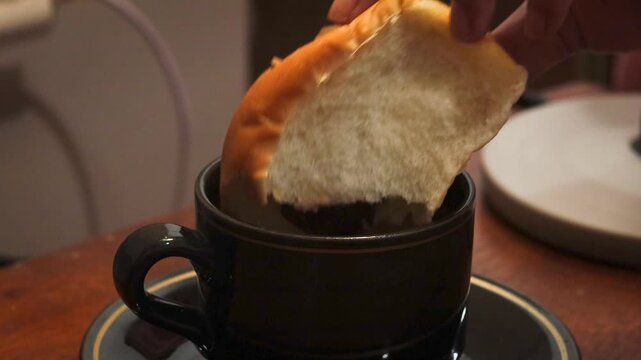 Asian woman's hand dipping a piece of bread into a cup of coffee at home. Simple breakfast and everyday dining activity.