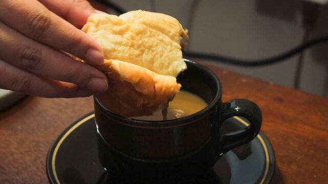 Asian woman's hand dipping a piece of bread into a cup of coffee at home. Simple breakfast and everyday dining activity.