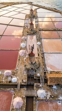Traditional stone windmills standing over geometric salt pans at the Sicily coastline with sunset sky reflections