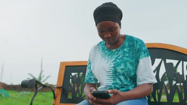Black woman using phone on bench with green field backdrop, wearing turquoise patterned shirt and headwrap, focused on screen while scrolling messages, relaxed posture and gentle smile, visible