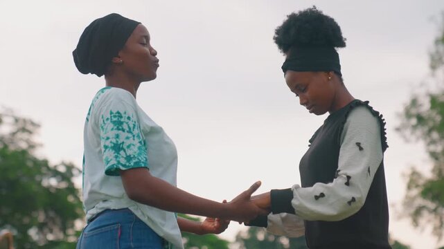 Black mother and daughter holding hands, quiet supportive moment in park at dusk side profiles, gentle eye contact, comforting gesture, casual clothing with headwrap and afro, soft muted sky