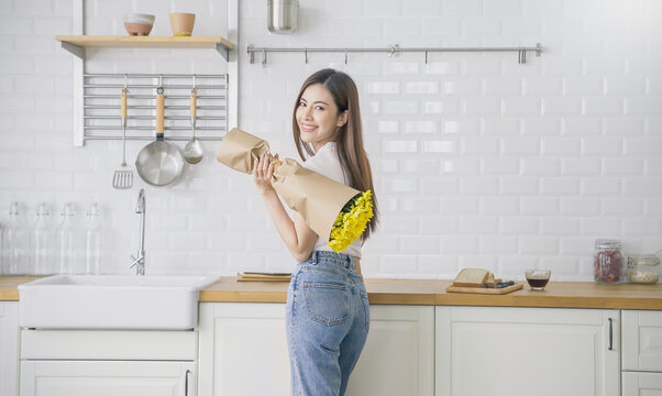Young asian woman stand at kitchen counter relaxing in her living room holding bouquet of yellow flowers at home. beautiful girl stay weekend minimal hobby or leisure lifestyle