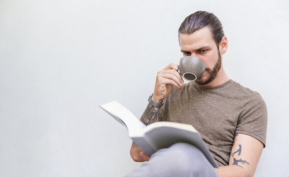 Young bearded man sitting on a couch at home reading a book, calm lifestyle moment with minimalist concrete wall background. Hobby leisure time authentic concept