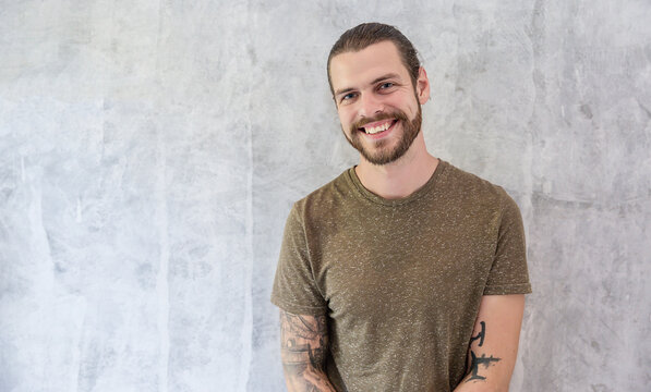 Portrait of bearded caucasian man with tattoos standing against a textured gray background, expressing confidence, friendliness, and a casual modern lifestyle.