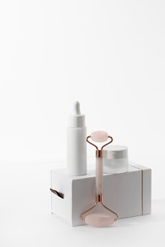 Minimalist facial care set featuring a rose quartz roller, white dropper bottle, and glass cream jar atop a sleek gift box. A clean, high-key studio shot perfect for luxury skincare branding.