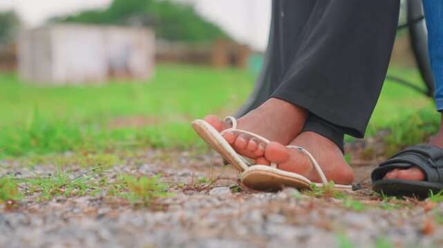 Mother and child feet close sitting on gravel yard with sandals, toes touching, lowangle closeup of adult and child legs, green grass and blurred village backdrop, quiet nurturing moment, caregiver