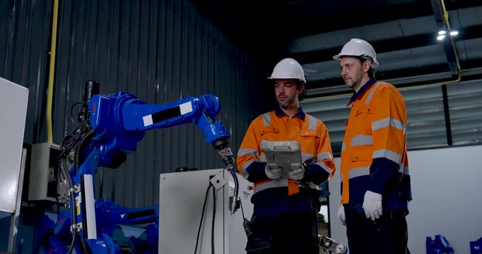 Two adult male technicians observe industrial robotic arm movement, reviewing handheld controller while monitoring automated machinery operation during manufacturing work in factory workshop.