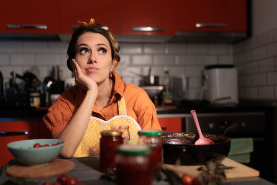 
Bored Housewife Waiting For Jam To Cook In Red Kitchen. Tired young woman leaning on her hand looking up thoughtfully while cooking