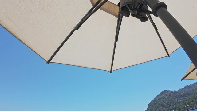 Under the umbrella by the pool at the hotel. Sunbath concept. Blue sky and beach umbrella 4k video. Summer vacation, resort hotel.