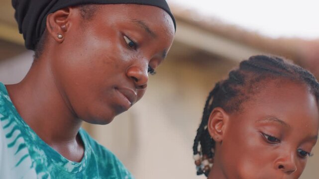 Black mother and child closeup bonding, mother watching daughter with tender gaze as child studies small object braided hair, warm natural light, quiet residential background, calm nurturing