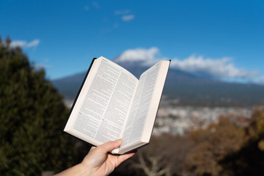 Outdoors, an open Bible showing Jeremiah chapter 9 is held by a hand against Mount Fuji and Fujinomiya city from Shirao Mountain Park (Shiraosan). Horizontal shot.