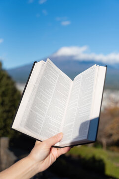 Outdoors, an open Bible showing Jeremiah chapter 9 is held by a hand against Mount Fuji and Fujinomiya city from Shirao Mountain Park (Shiraosan). Vertical shot.