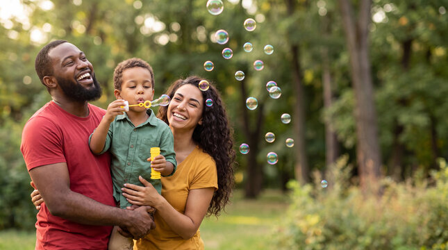 Family of three enjoying a sunny day outdoors, father holding son while mother smiles, bubbles floating in the air around them
