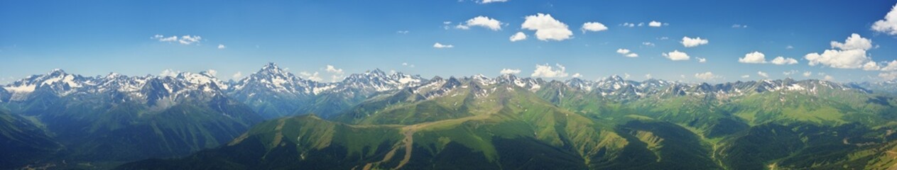 Fototapeta premium Extra wide panoramic landscape of a majestic mountain ridge with snowy peaks, glacier ice and green forested valleys under a blue sky with day haze.