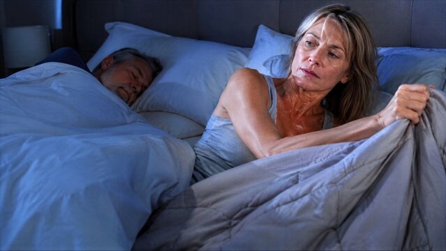 Sudden menopause heat wakes mature woman beside sleeping partner, while one simple movement of blanket reveals different temperature needs and broken sleep at night at home.