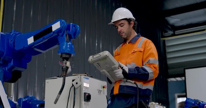 Adult male technician controls industrial robotic arm with handheld pendant in factory workshop, looking between controller and machinery while testing automated movement and equipment response.