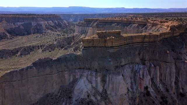 Aerial drone footage captures Utah Scenic Byway 12 region with dramatic layered canyon walls and sedimentary rock formations. Vast desert landscape stretches toward distant mesas under hazy blue sky.