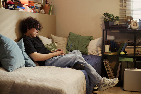 Asian young man sitting on bed and typing on laptop computer, studying or working from home. Image represents remote work, online education, and digital lifestyle, wide shot