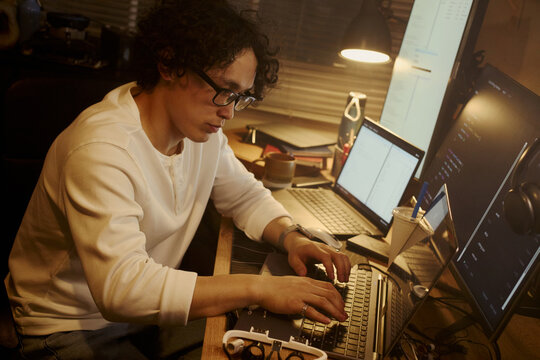 Asian young man typing on laptop at desk with multiple screens, coding software at night. Image represents remote work and IT development, suitable for tech recruitment ads