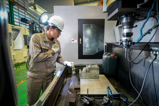 CNC machine machinist factory manufacturing metal safety hardhat glove, focused male worker operate milling equipment inside workshop, inspecting workpiece and tool under bright task light
