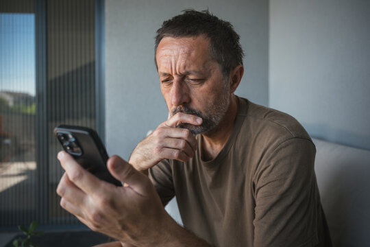 Middle-aged man sits on a balcony sofa near, intently looking at his smartphone with a worried expression, conveying focus, solitude and the everyday tension of modern digital life.