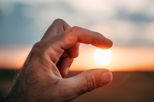 Close-up of a human hand pinching the setting sun between two fingers against a colorful evening sky. Creativity, perspective and playful nature concept.