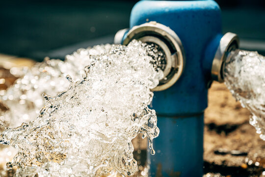 Action shot of clean water gushing and spraying intensely from a bright blue outdoor utility hydrant in the summer.