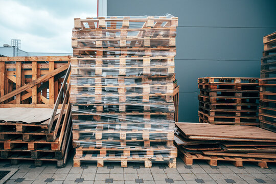 Stack of used wooden euro pallets wrapped in transparent stretch film in front of a warehouse building. Logistics, shipping and industrial storage concept.