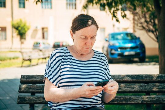 Middle-aged woman enjoying a quiet moment while checking her smartphone in a leafy city park. Digital communication, relaxation and modern everyday life theme.