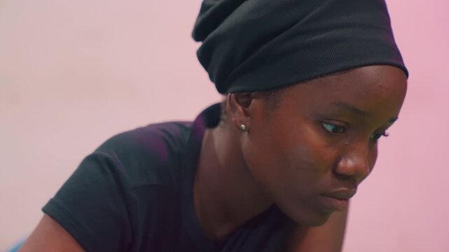 Young black mother looking down contemplative, headscarf closeup with soft pink background, subtle sweat on brow, quiet fatigue, introspective mood, caregiver reflection and resilient dignity