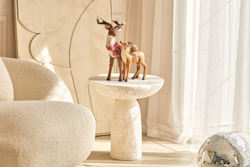 Christmas reindeer figurines decorate a side table in a cozy home interior, with warm sunlight, neutral beige decor, sheer curtains, armchair and festive silver ornament creating a holiday scene. © Yulia Mihaylova
