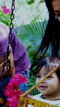 Indian Asian young parents gently swinging twin infant babies on playground swing in outdoor park, smiling as babies laugh and enjoy fresh air, nature greenery and joyful family bonding time together