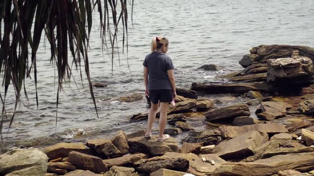 Asian female carefully navigates the rugged rocks along the coastline of Khao Laem Ya Mu Ko Samet National Park in Thailand, framed by overhanging tropical foliage