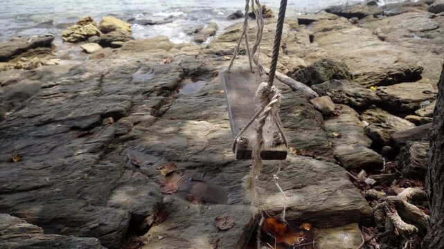 An empty rustic wooden rope swing sways gently in the ocean breeze above the rocky shoreline at Khao Laem Ya Mu Ko Samet National Park, offering a tranquil seaside escape