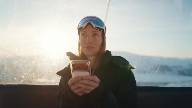 Female travel blogger posting in social media by smartphone, riding cable car. Portrait of beautiful woman in activewear and sunglasses for snowboarding or skiing chatting online in mobile app