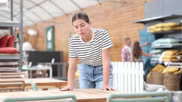 Girl customer choose dining table desk in garden furniture store showroom center. Young woman runs hand over smooth surface, imagining how it would look in patio 
