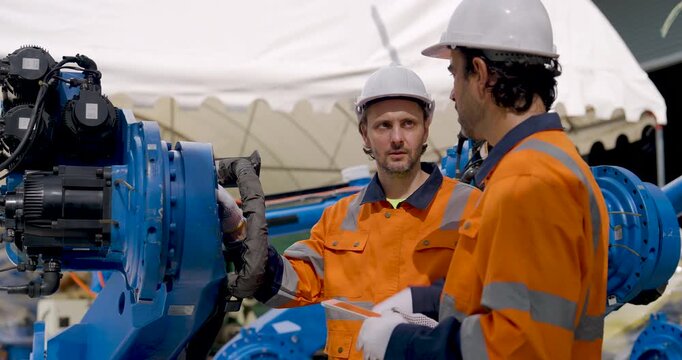 Two adult male technicians discuss industrial robotic arm condition beside factory equipment, using tablet and gestures while planning inspection and maintenance for automated manufacturing.