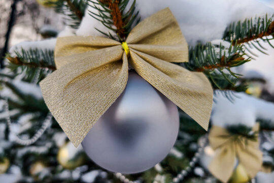 A natural Christmas tree stands outside in winter with decorations. A silver ornament hangs from a branch secured by a golden bow