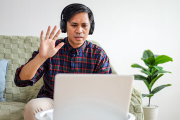 Asian Man Wearing Headphones Waving Hand During laptop Video Call at Home © Gatot