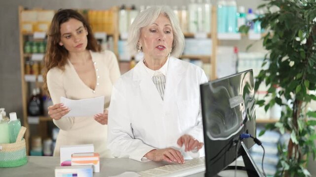 Senior woman employee in labcoat works in pharmacy using computer and collaborate with female client. Professional comment doctors prescription, recommend drug for treatment regiment 