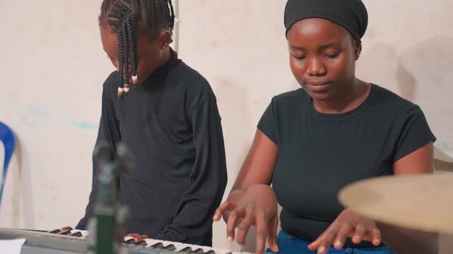 Mother coaching child keyboard in rehearsal, quiet worshipful practice with drums and microphone in foreground, focused mentor leading young musician through hymn and harmony, calm devotional