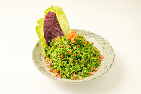Tabbouleh salad with pomegranate seeds and diced tomato, garnished with cabbage leaves in a textured bowl.