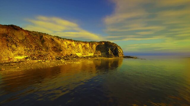 Golden cliffs and calm sea shore, sunlit limestone face glowing against tranquil water, soft clouds stretching to distant horizon, pebbled foreground