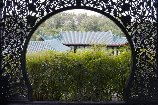 A serene view of a traditional building with grey tiled roofs and curved eaves, framed by an intricate circular metal gate in GuiHu Park, Chengdu. Lush green foliage creates a natural layer between