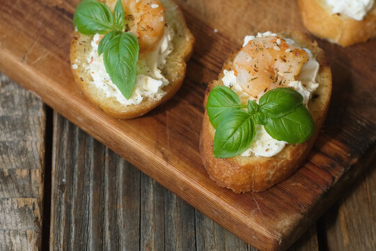 Tasty snacks. Bruschetta or crostini. Toasted baguette slices with feta cream cheese, shrimp and fresh basil on wooden board. 