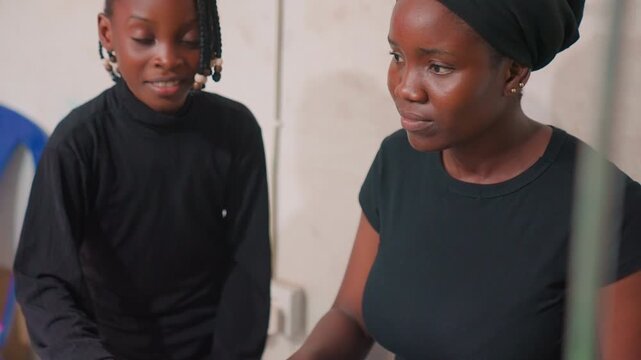 Black mother and child seated in modest home, mother guiding daughter through quiet conversation, closeup interaction, gentle expressions, knitted headscarf, casual black clothing, soft daylight,