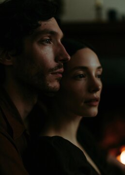 Close-up portrait of a man and a woman. the man is on the left side of the image, with dark hair and a beard. he is looking off to the side with a serious expression on his face.