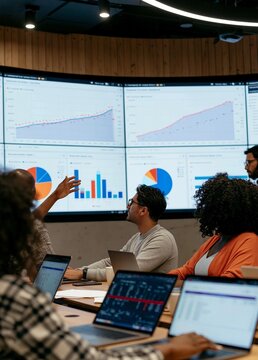 Group of people sitting around a table with laptops in front of them. they are in a conference room with large screens on the wall displaying graphs and charts.